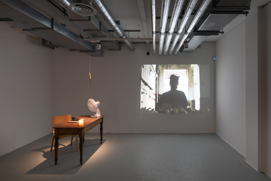 Small dark gallery space with projected image on rear wall and a lit table with artworks